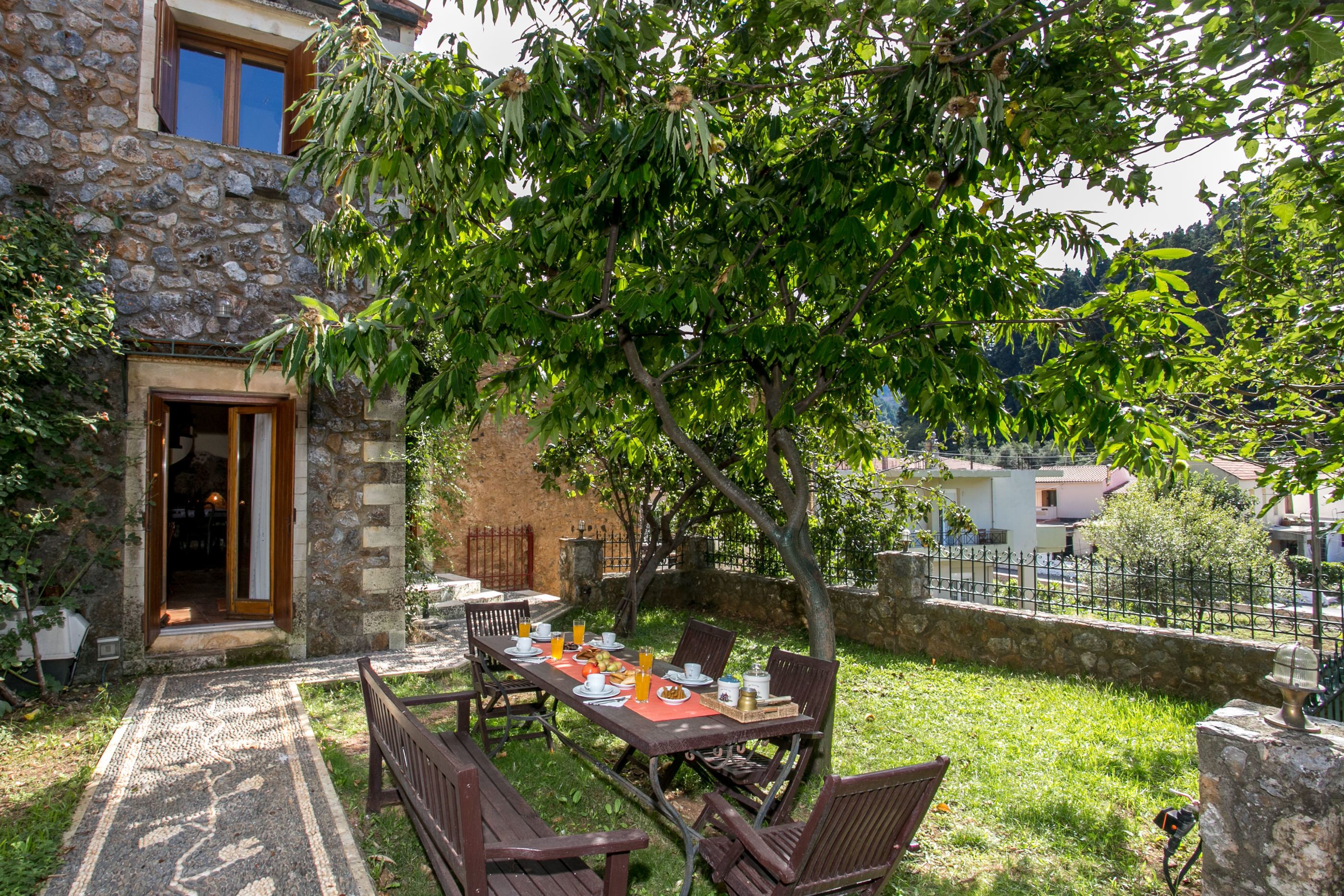 An outdoor dining area with a stone building, a table set for two, and a tree providing shade.