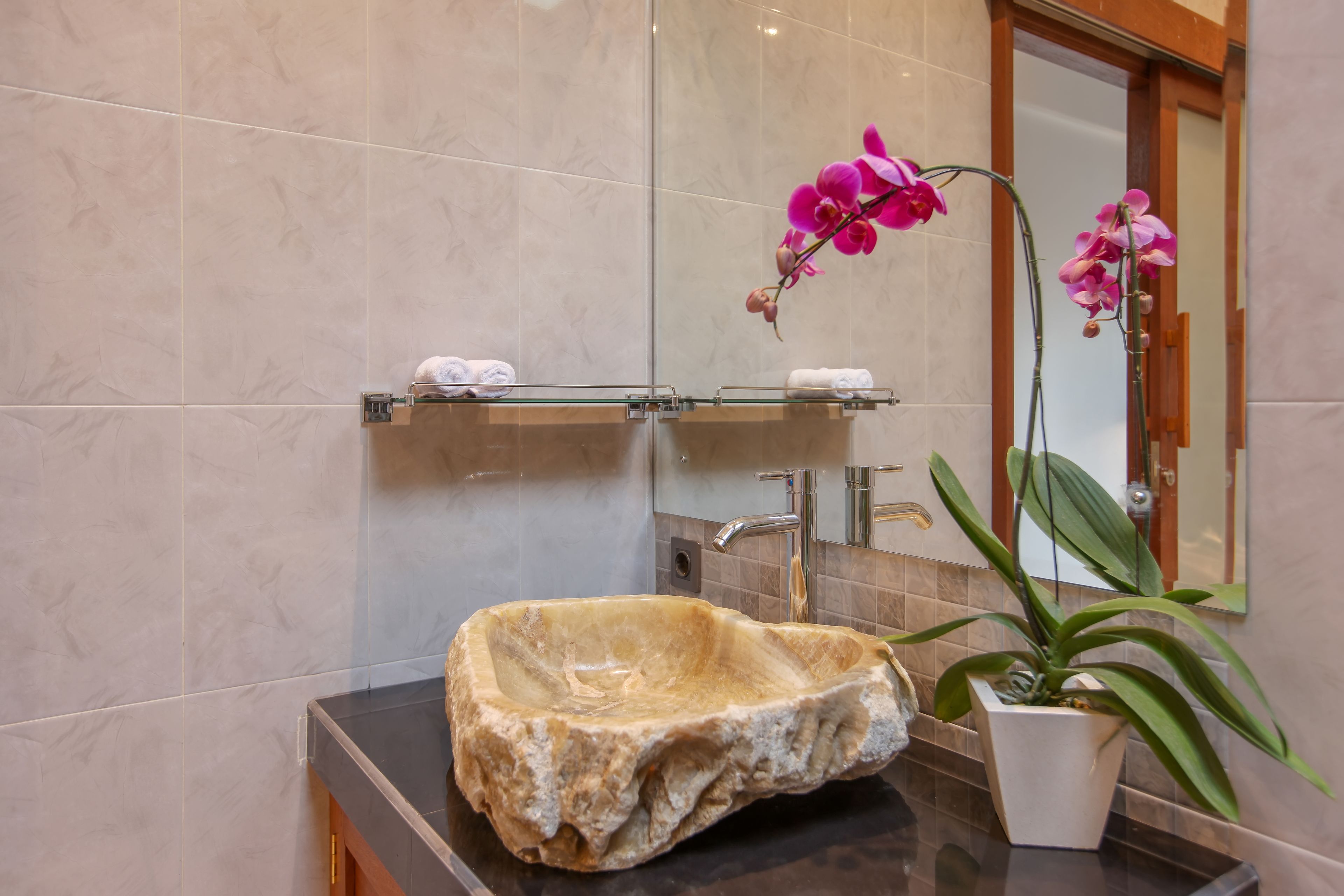 Un baño con un lavabo de piedra, una repisa de vidrio con toallas, un espejo y una orquídea en maceta.
