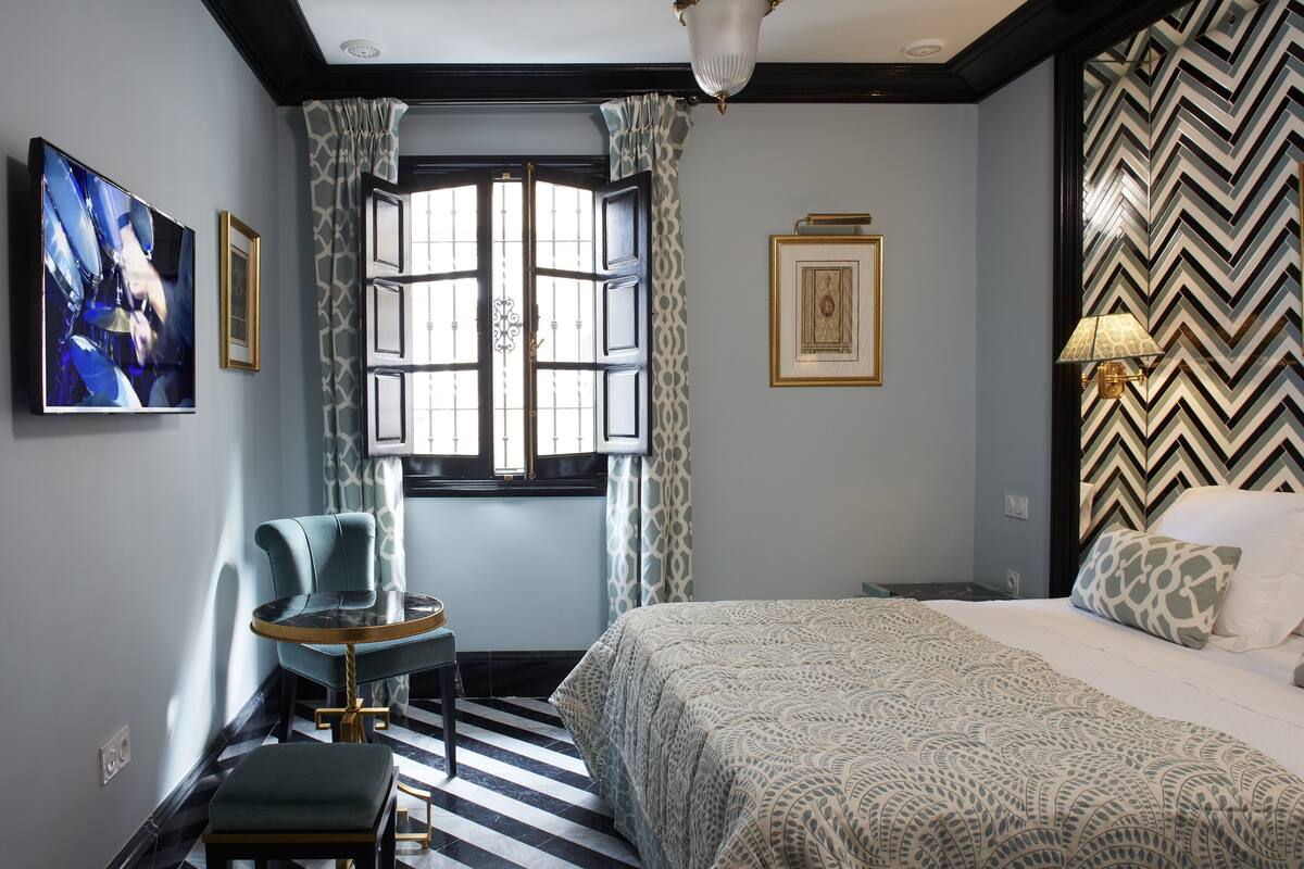 Une chambre d’hôtel avec un lit, une chaise, une petite table, une fenêtre avec des rideaux et un mur décoré de motifs.