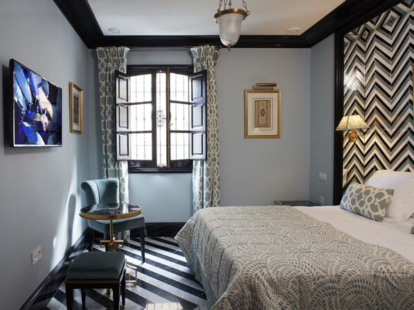A hotel room with a bed, a chair, a small table, a window with curtains, a wall-mounted picture, and a patterned wall. - Triana House (Seville)