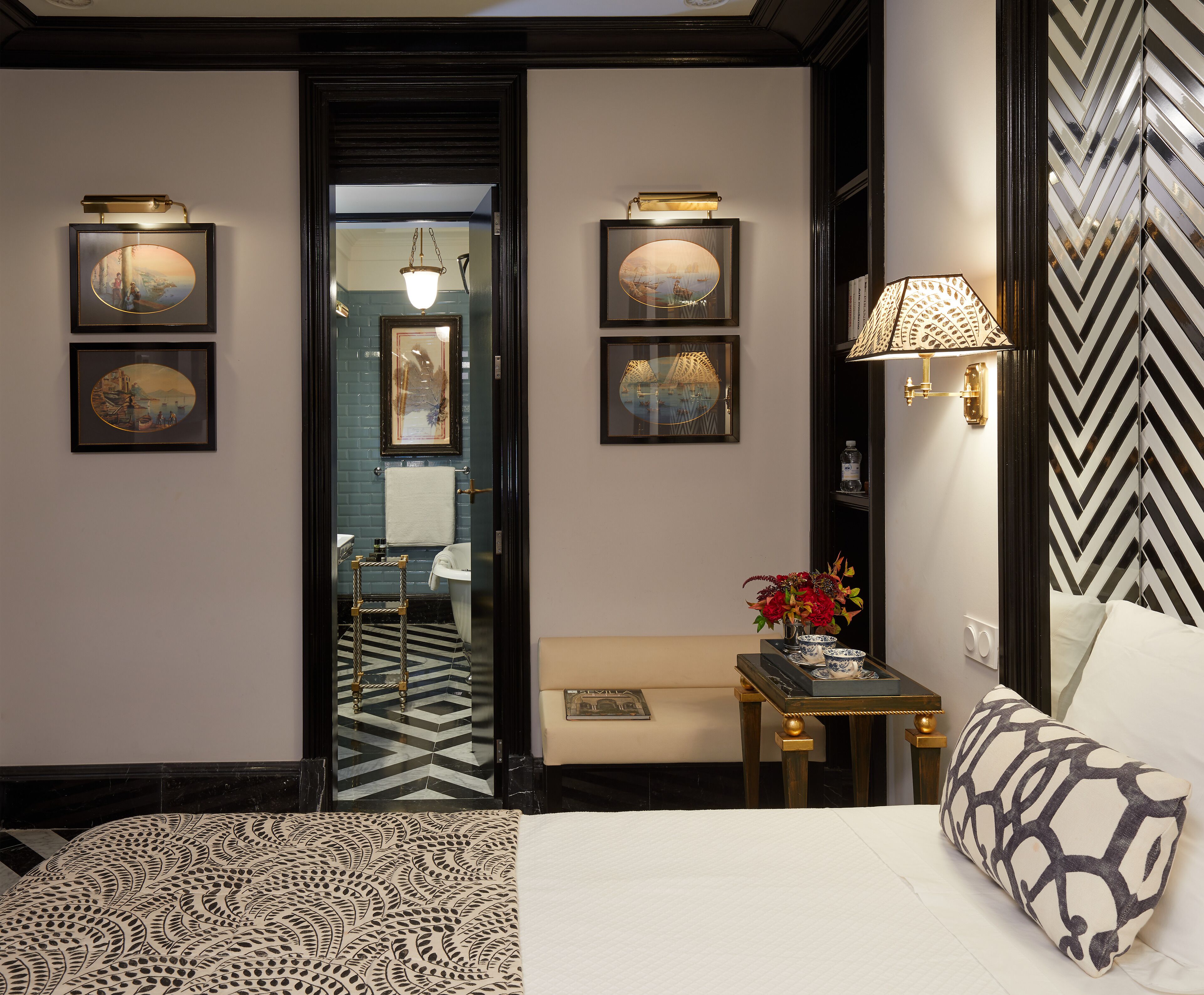 A hotel room with a bed, a bench, a small table with a vase of flowers, and framed pictures on the walls.