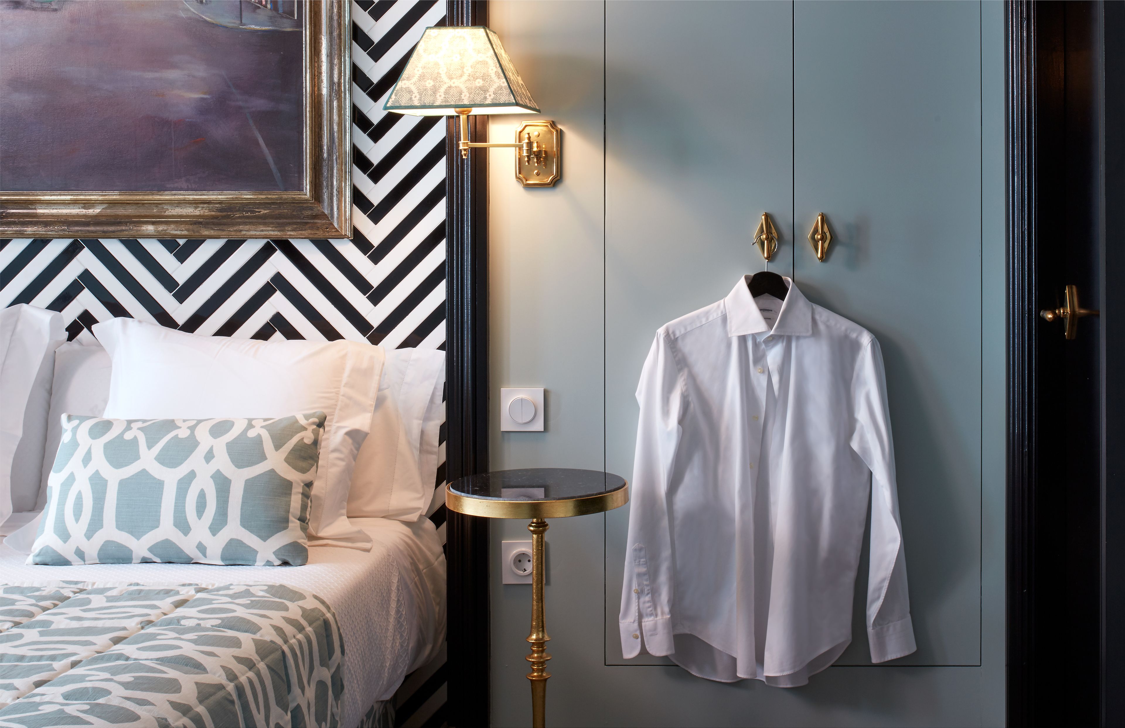 A neatly arranged bedroom with a bed, pillows, a bedside table, a lamp, and a hanging shirt.