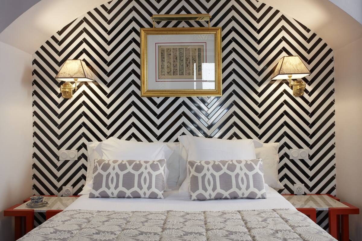 Une chambre à coucher bien aménagée, avec un papier peint à motifs chevrons, un tableau encadré au-dessus du lit et deux lampes de chevet.