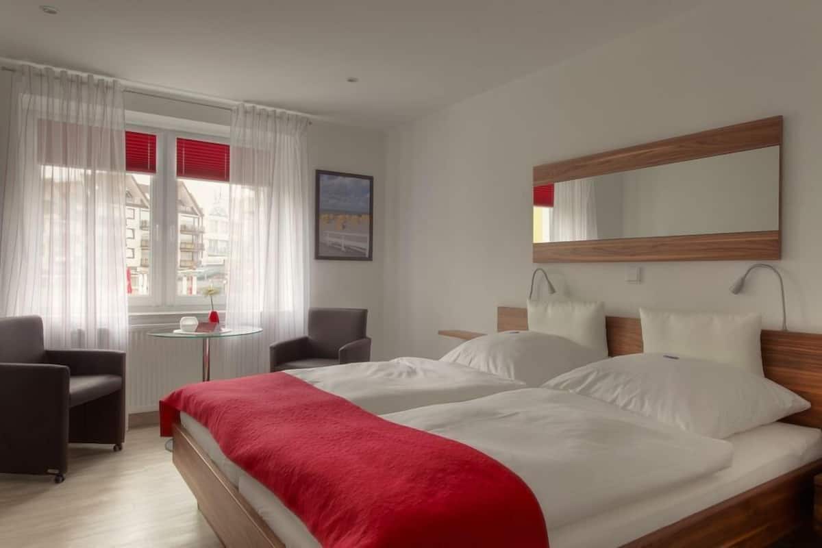 A hotel room with a large bed, two bedside tables, a chair, a small round table, and a window with curtains.