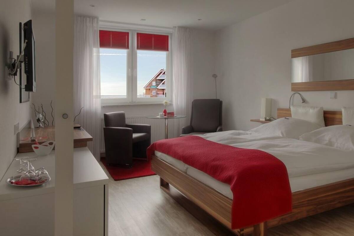 A modern hotel room with a large bed, a desk, a chair, and a view of buildings through the window.