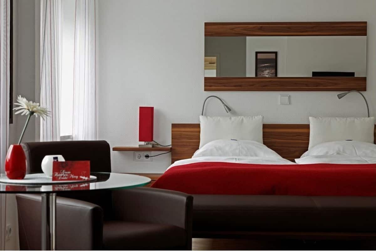 A hotel room with a bed, a bedside table, a red vase, a glass table, and a chair.