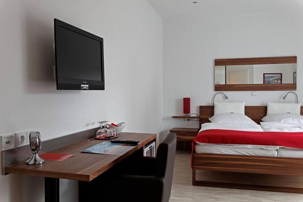 A hotel room with a bed, a desk with a chair, a TV mounted on the wall, and a bedside table with a lamp.