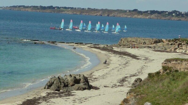 Une plage bordée d’une rangée de voiliers sur l’eau, un rivage rocheux et une vue lointaine sur des maisons.