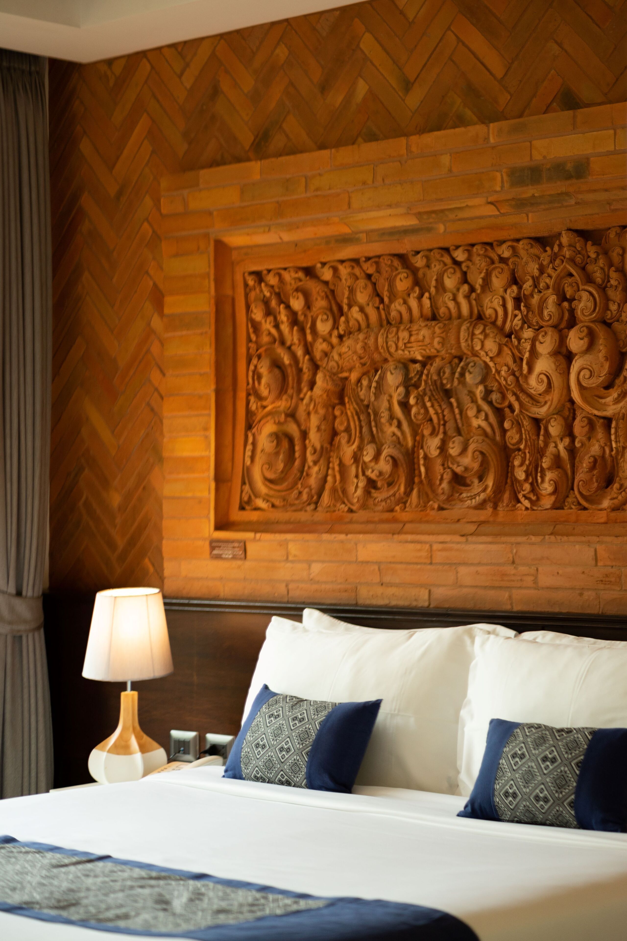 A hotel room with a herringbone-patterned wooden wall, a decorative carved panel above the bed, and a white bed with blue and patterned pillows.