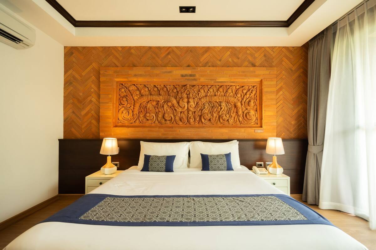 Une chambre d’hôtel avec un grand lit, deux tables de chevet avec des lampes, une tête de lit décorative en bois et une couvre-lit à motifs.