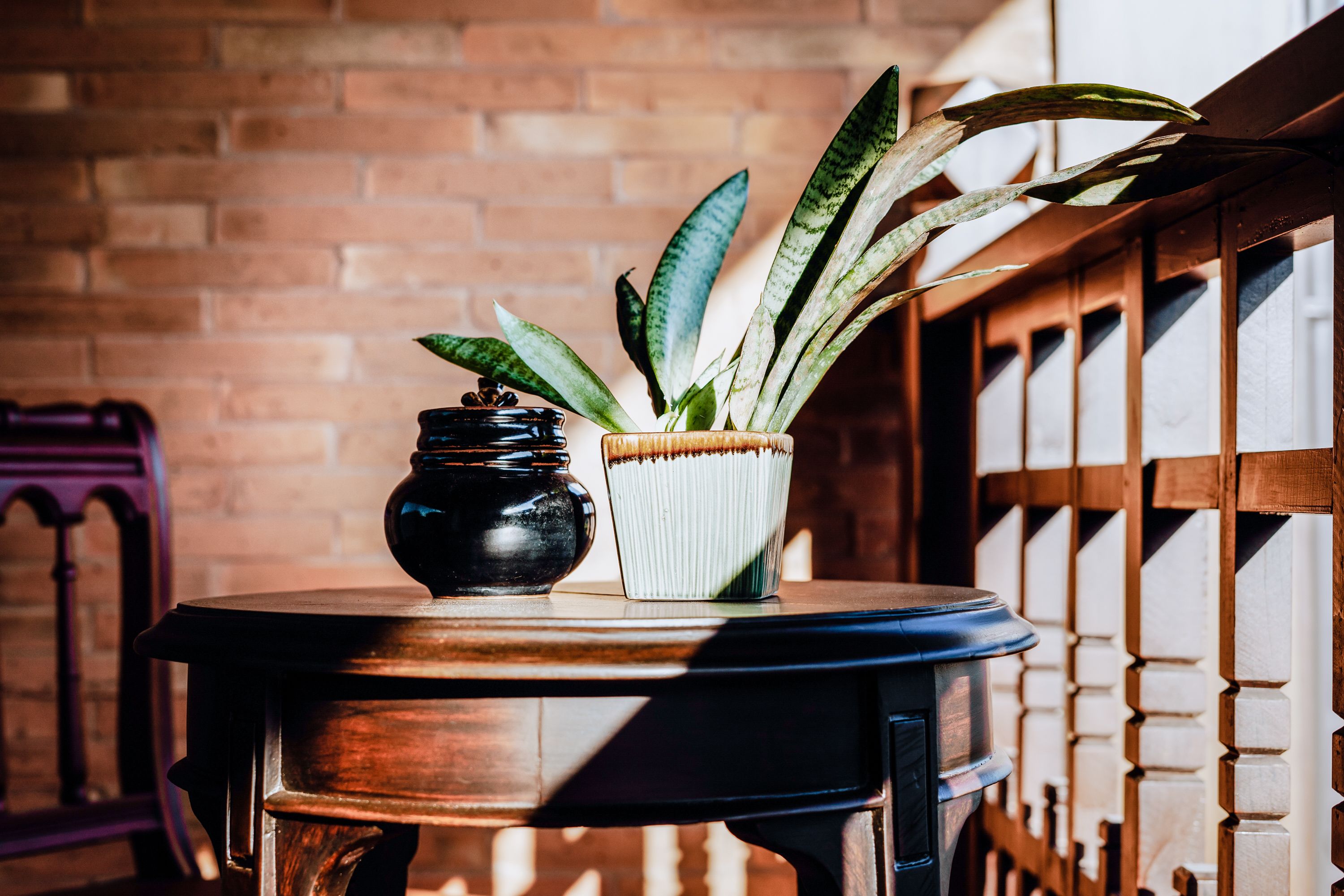 A round wooden table with two potted plants, one in a black pot and another in a striped pot, against a brick wall.