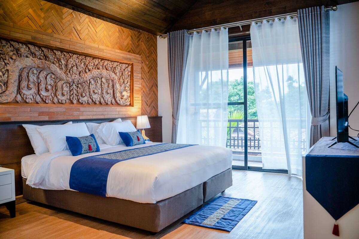 Une chambre avec un grand lit, une tête de lit en bois, un élément décoratif mural, une télévision et une vue sur l’extérieur par une fenêtre.