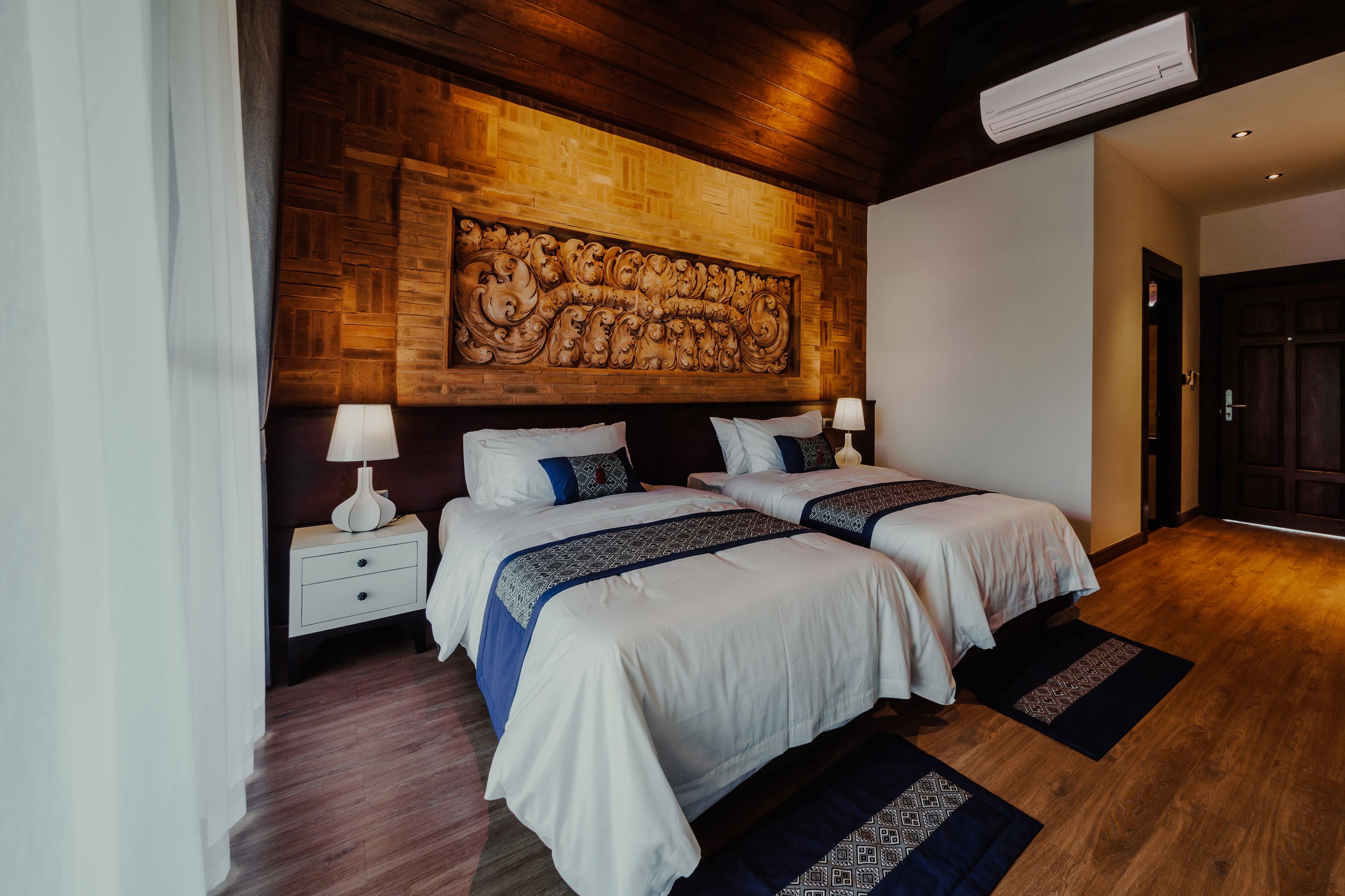 Camera d'albergo con due letti, una testiera in legno, un comodino con una lampada e un grande elemento decorativo a parete.