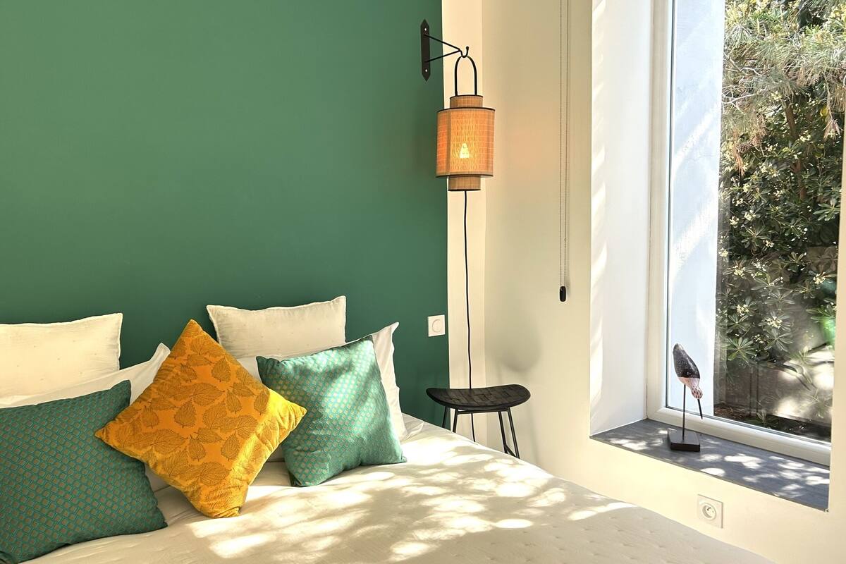 Ein Bett mit weißer Bettwäsche und mehreren Kissen, eine Stehlampe und ein Fenster mit Blick auf Grünflächen.