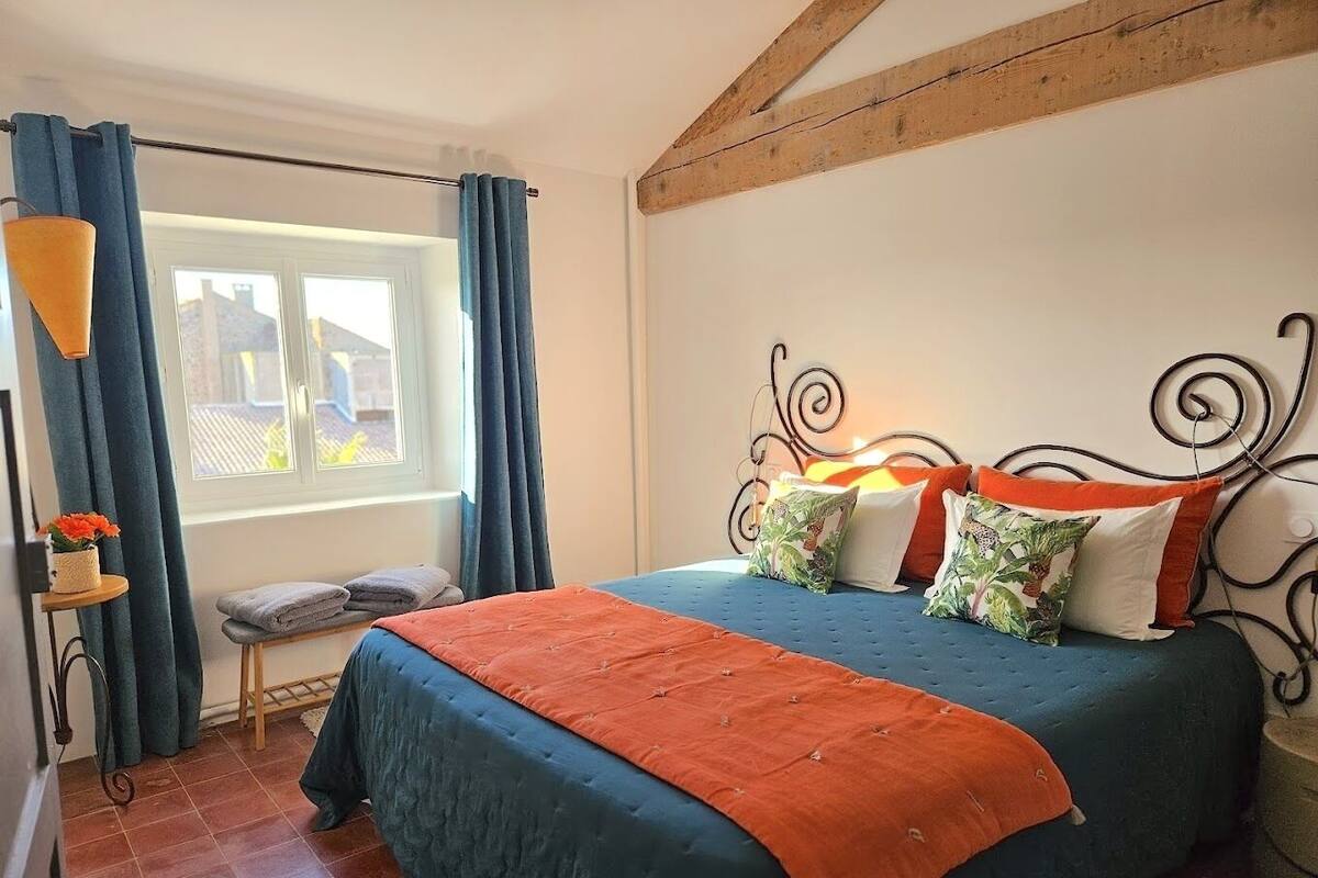 Ein Schlafzimmer mit einem Bett, Vorhängen, einem Fenster, einem Nachttisch und einer Wand mit sichtbaren Balken.