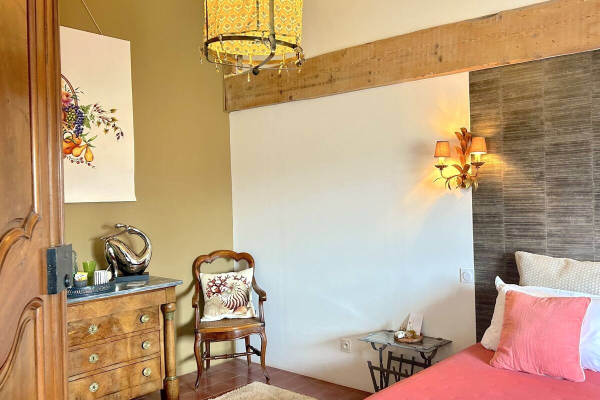 Ein Schlafzimmer mit Holzboden, einem Bett mit rosa Bettwäsche, einem Sessel mit Blumenkissen, einer Kommode mit Schubladen und einer Wandlampe.
