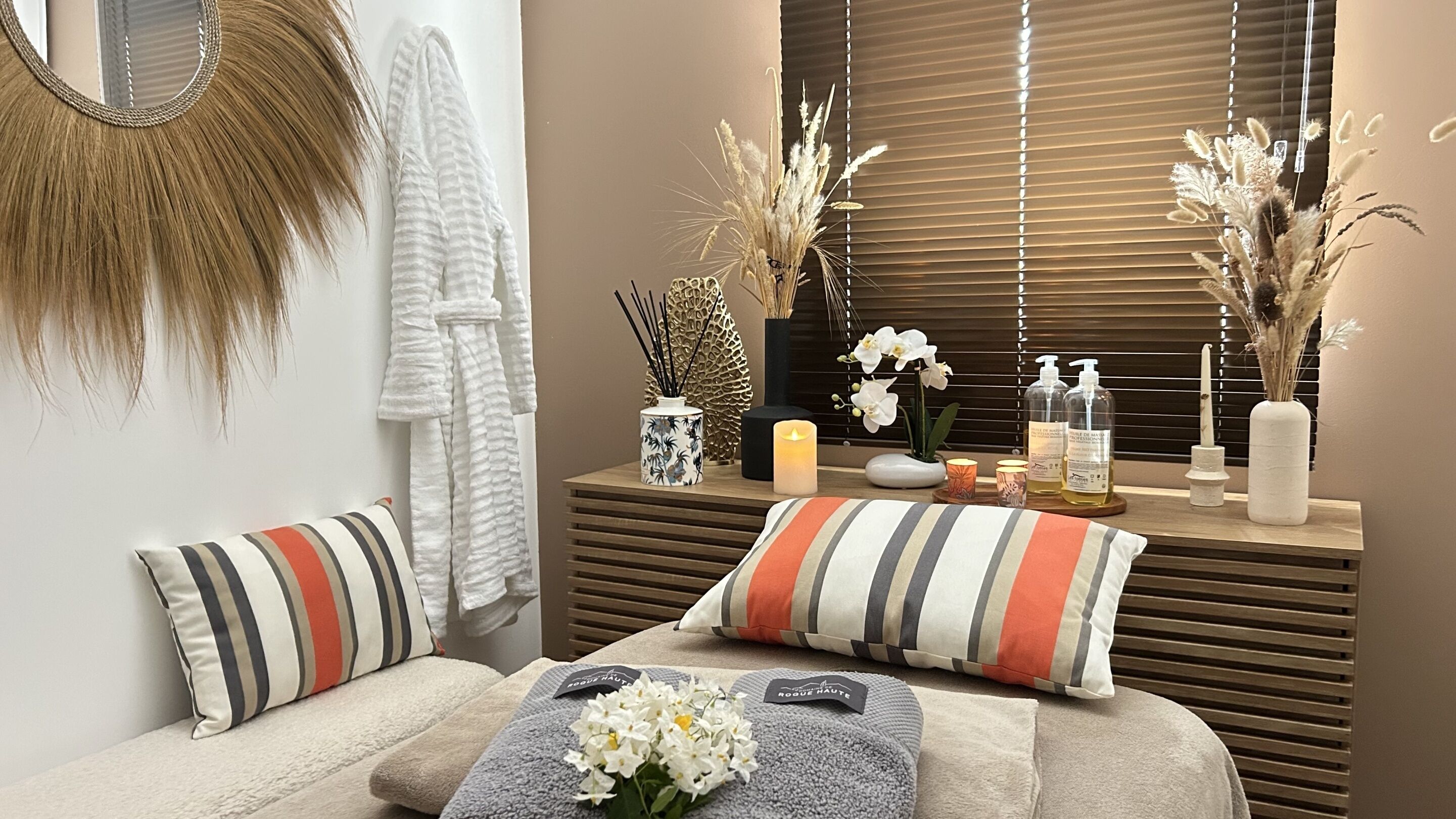A spa room with a massage table, striped pillows, a mirror, a vase with dried plants, candles, and a bottle of lotion.
