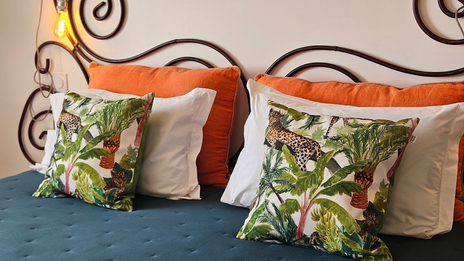 Un letto con due cuscini decorativi, uno con una fantasia giungla e l'altro con un motivo leopardato, e un cuscino arancione su una testiera imbottita blu.