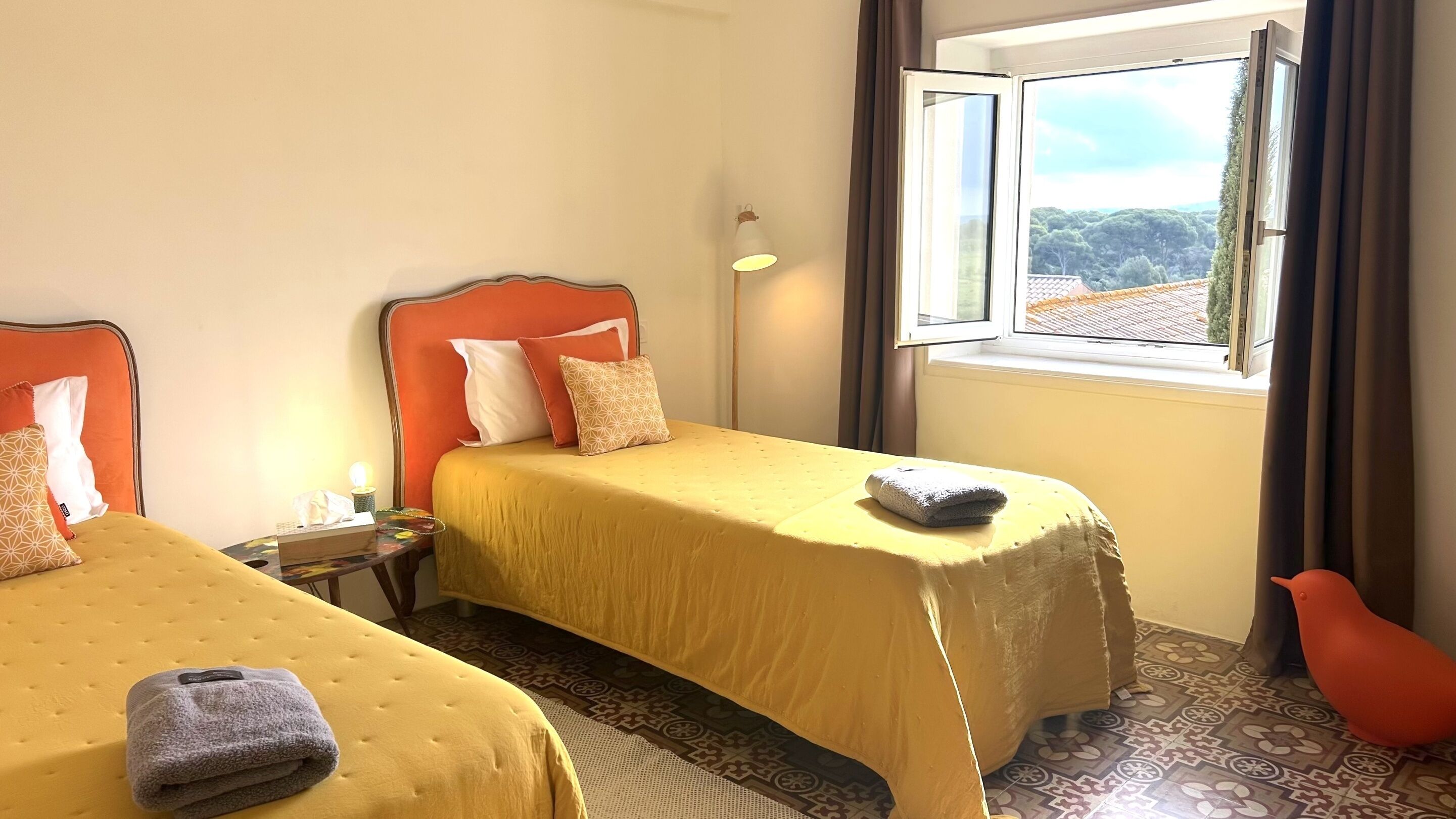 A hotel room with two beds, a nightstand, a lamp, and a window with a view.