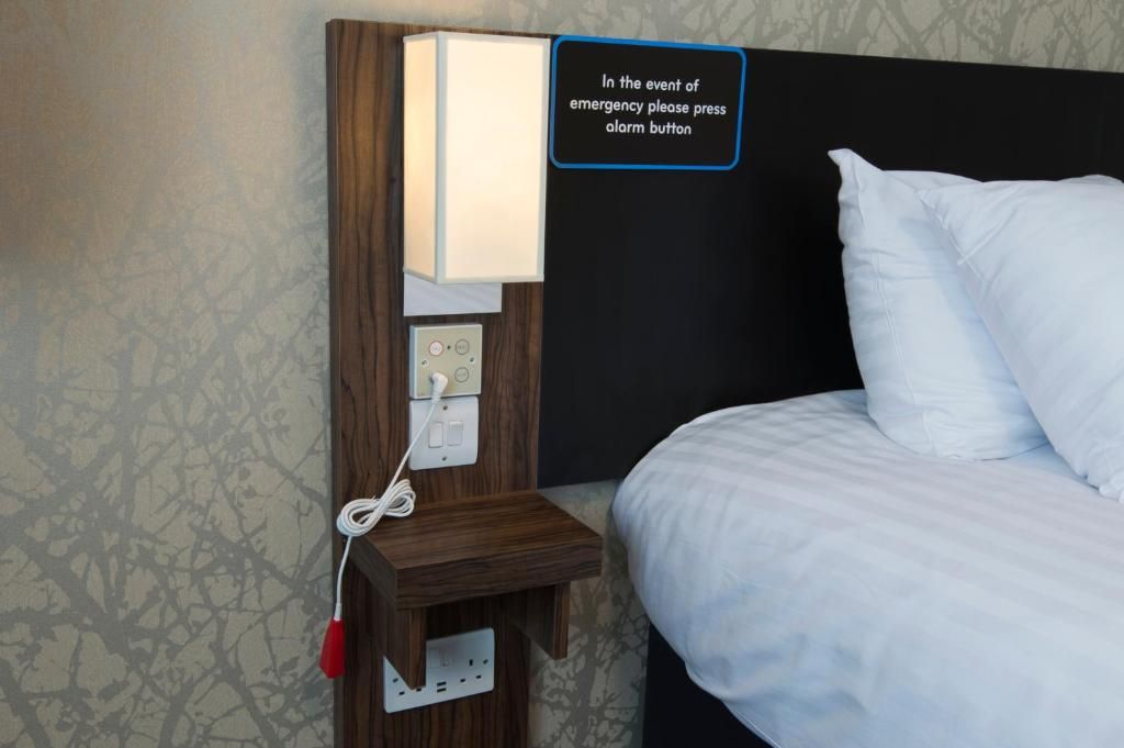 Una habitación de hotel con mesita de noche, lámpara, botón de alarma y cama.