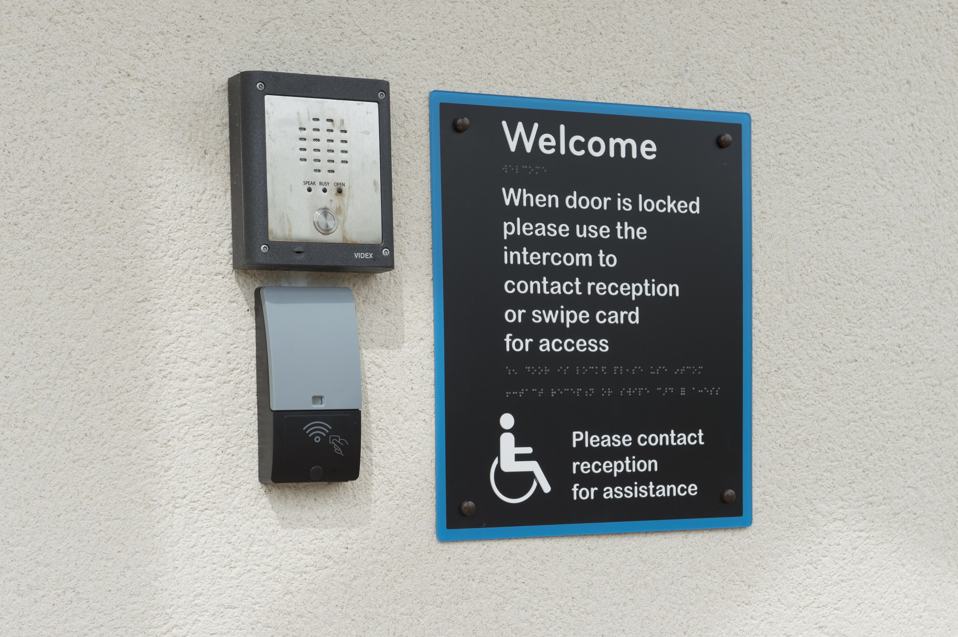 A wall-mounted intercom and a welcome sign with instructions for accessing a locked door.