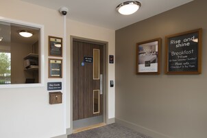 Zona de recepción de un hotel con puerta de madera, devolución de tarjetas de llave, carteles informativos enmarcados y una cámara de seguridad.
