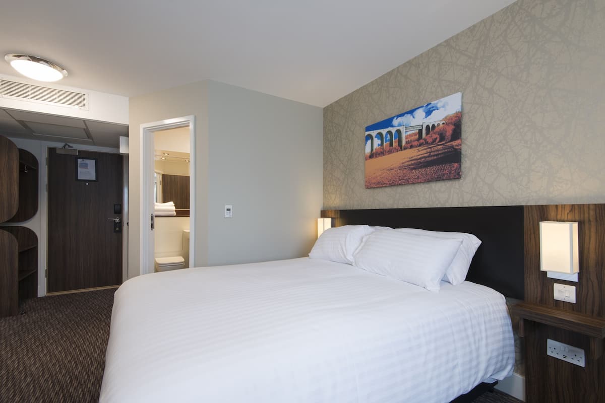 Une chambre d’hôtel avec un grand lit, une tête de lit en bois, un tableau accroché au mur et une porte donnant sur la salle de bain.