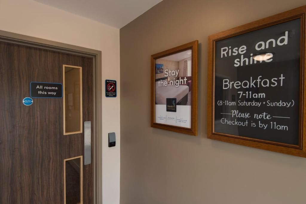 A hotel room with a sign indicating all rooms are this way, a framed picture with the text 'Stay the night,' and a sign for breakfast from 7-11am with a note that checkout is by 11am.