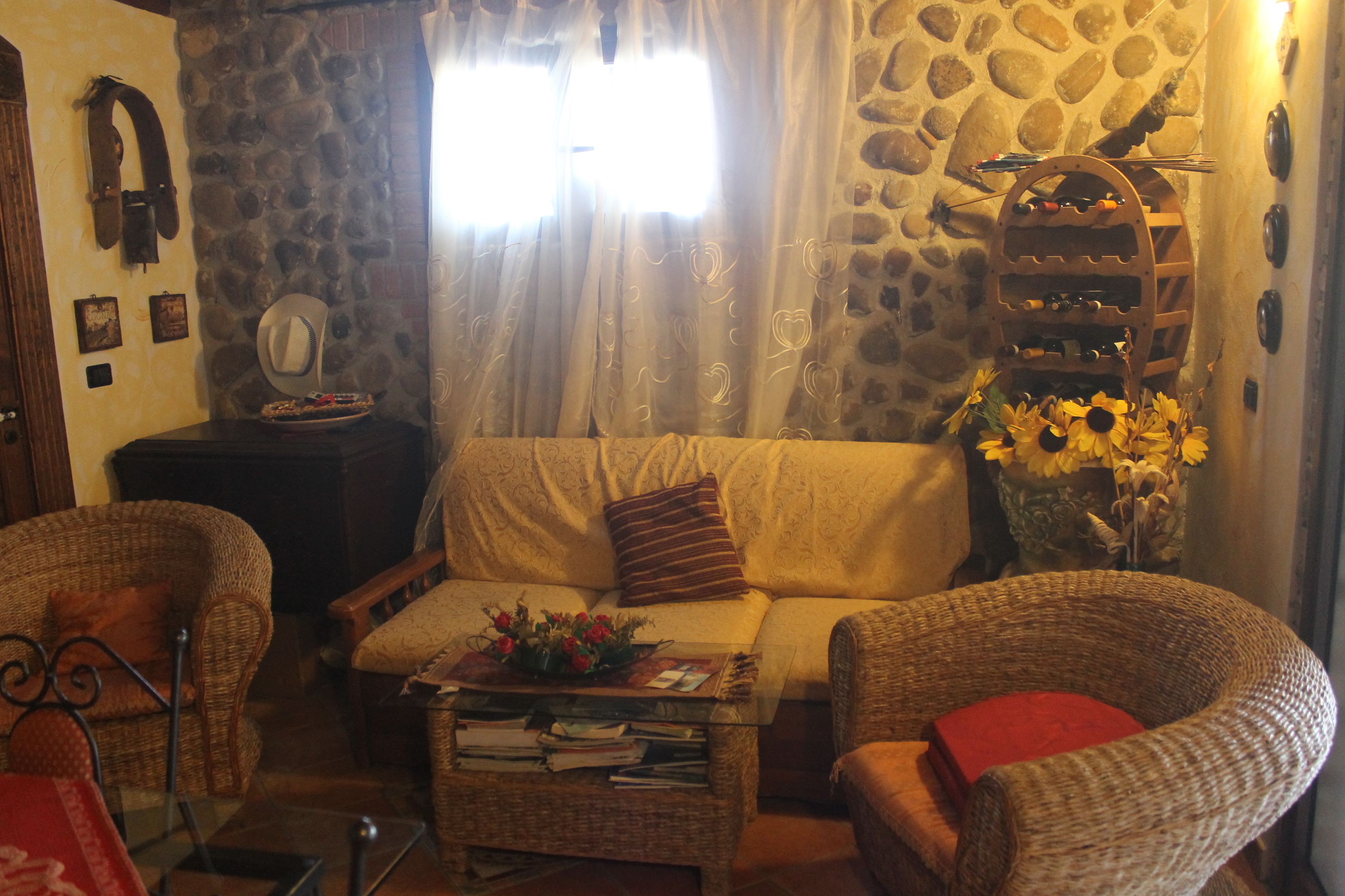 A cozy living room with a stone wall, a sofa, a coffee table with books and flowers, and wicker chairs.