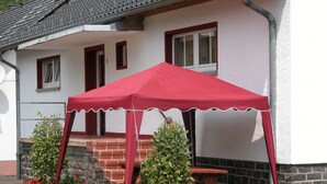 Ein rotes Zelt ist vor einem Gebäude aufgebaut, das eine Backstiege und einen Holztisch hat. Vor dem Zelt stehen Töpfe mit roten Blumen.