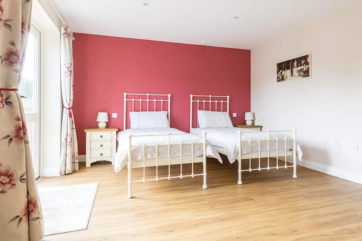 A bedroom with two white metal beds, a wooden floor, a red wall, and a window with curtains.