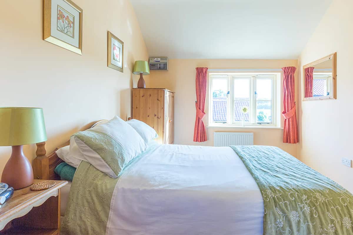 A bedroom with a bed, a nightstand, a lamp, a wooden cabinet, a window with curtains, and framed pictures on the wall.