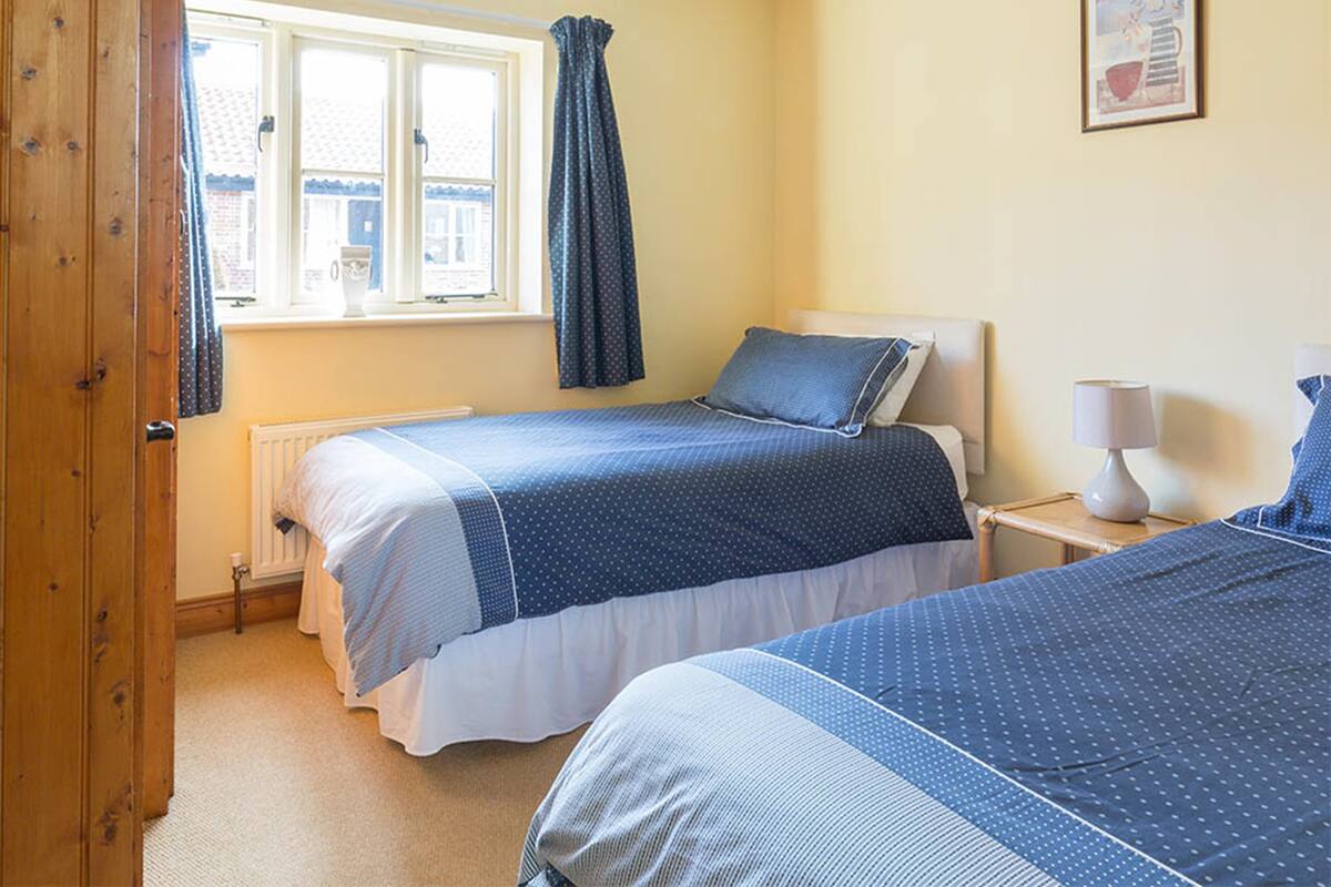 A bedroom with two beds, a window with curtains, a bedside table with a lamp, and a framed picture on the wall.