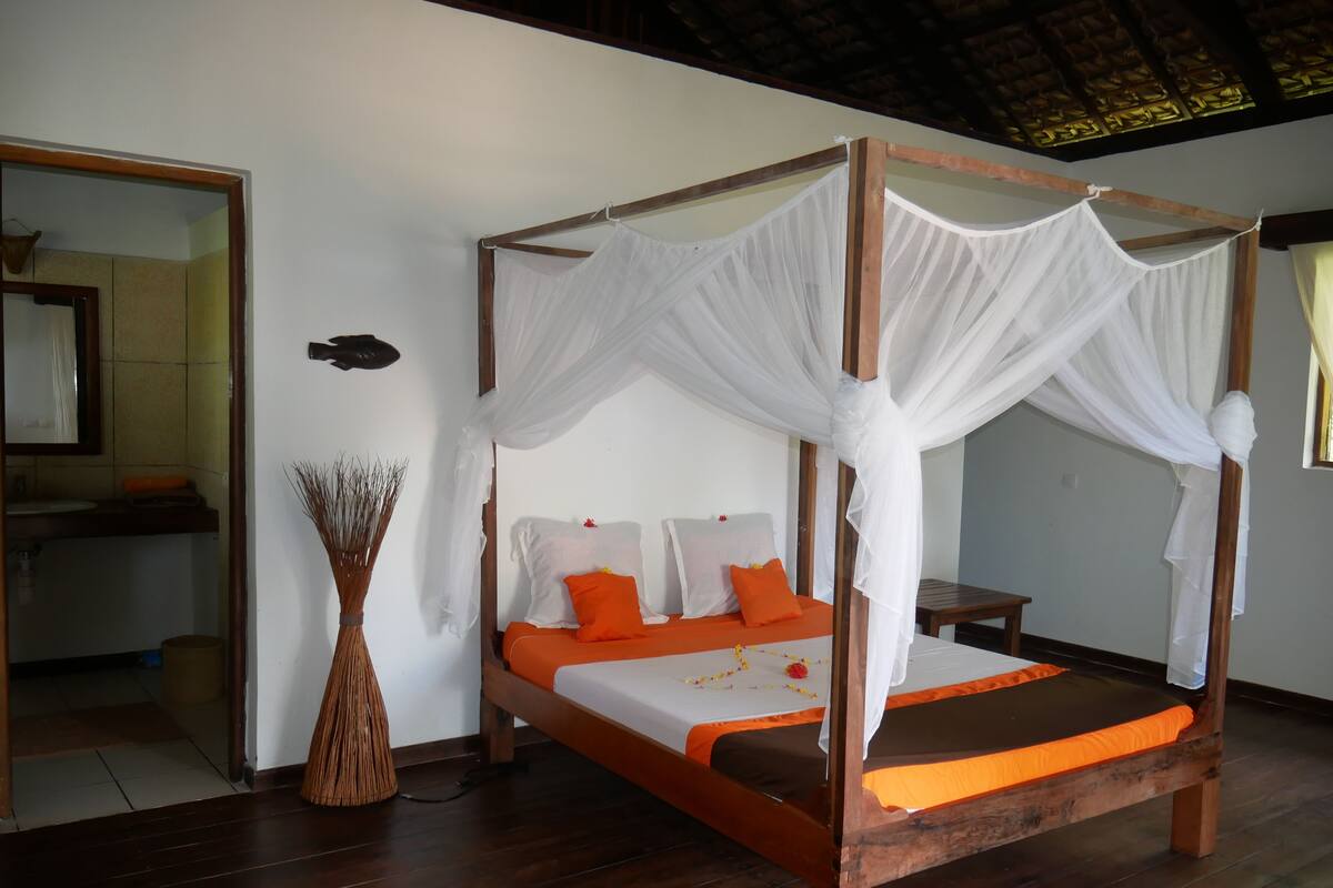 A four-poster bed with white drapes, orange bedding, and a wooden floor.