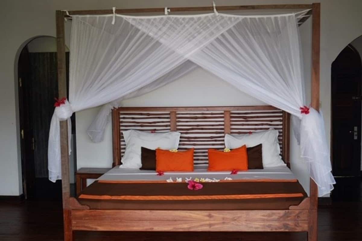 A four-poster bed with white canopy and wooden headboard, orange and white pillows, and a wooden floor.