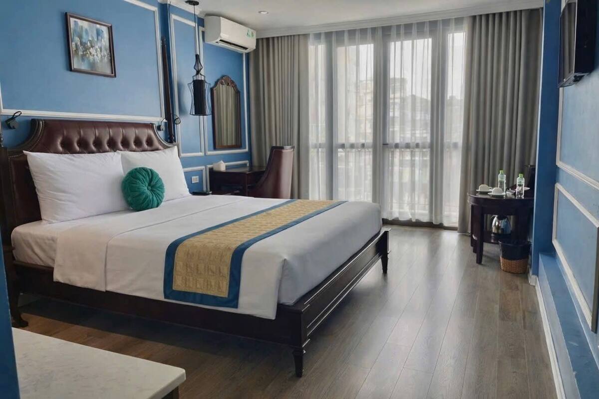 A hotel room with a large bed, a bedside table, a chair, a window with curtains, and a view of the city.