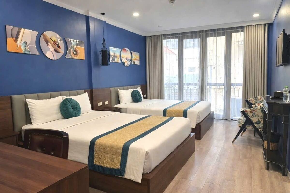 A hotel room with two beds, a desk, and a chair. There are large windows with curtains, artwork on the walls, and a balcony visible through the windows.