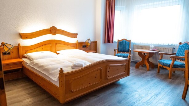 A wooden bed with a headboard, two bedside tables, a small round table, two chairs, and a window with curtains. - Röhrs Gasthof (Sottrum)