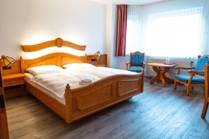 Ein Bett mit Holzkopf, zwei Nachttische, ein kleiner runder Tisch, zwei Stühle und ein Fenster mit Vorhängen.
