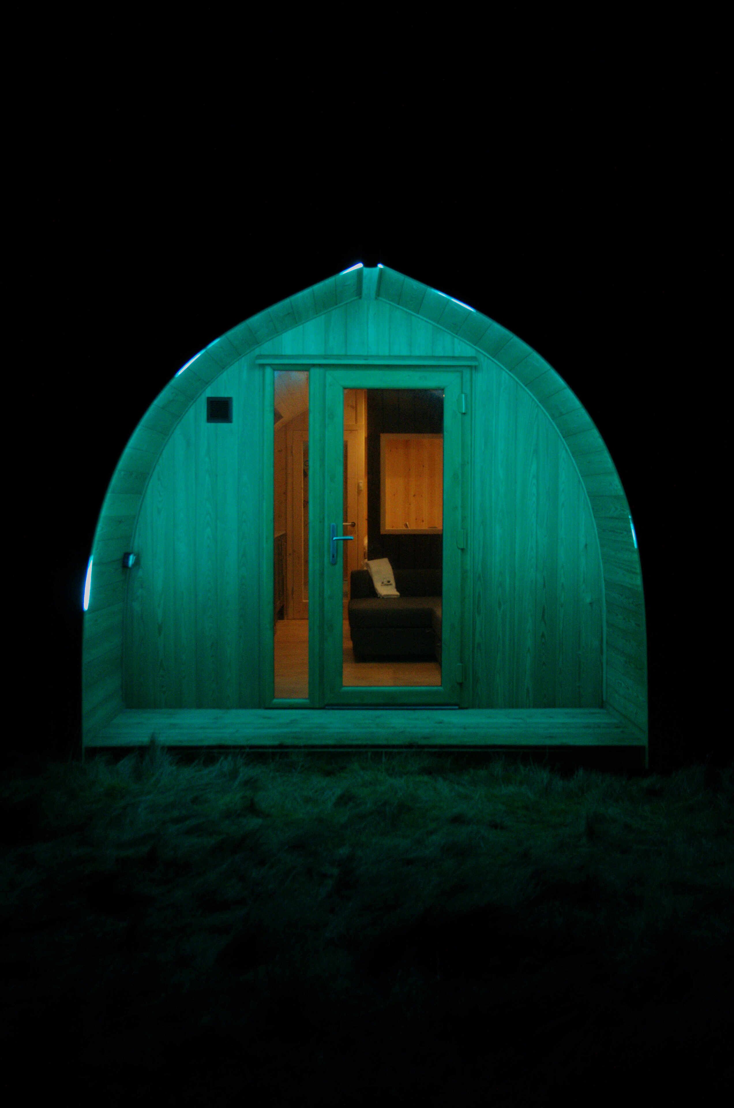 A small, single-room structure with a curved roof, a door leading inside, and a visible interior with a bed and a small table.