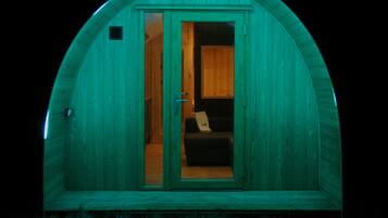 A small, single-room structure with a curved roof, a door leading inside, and a visible interior with a bed and a small table.