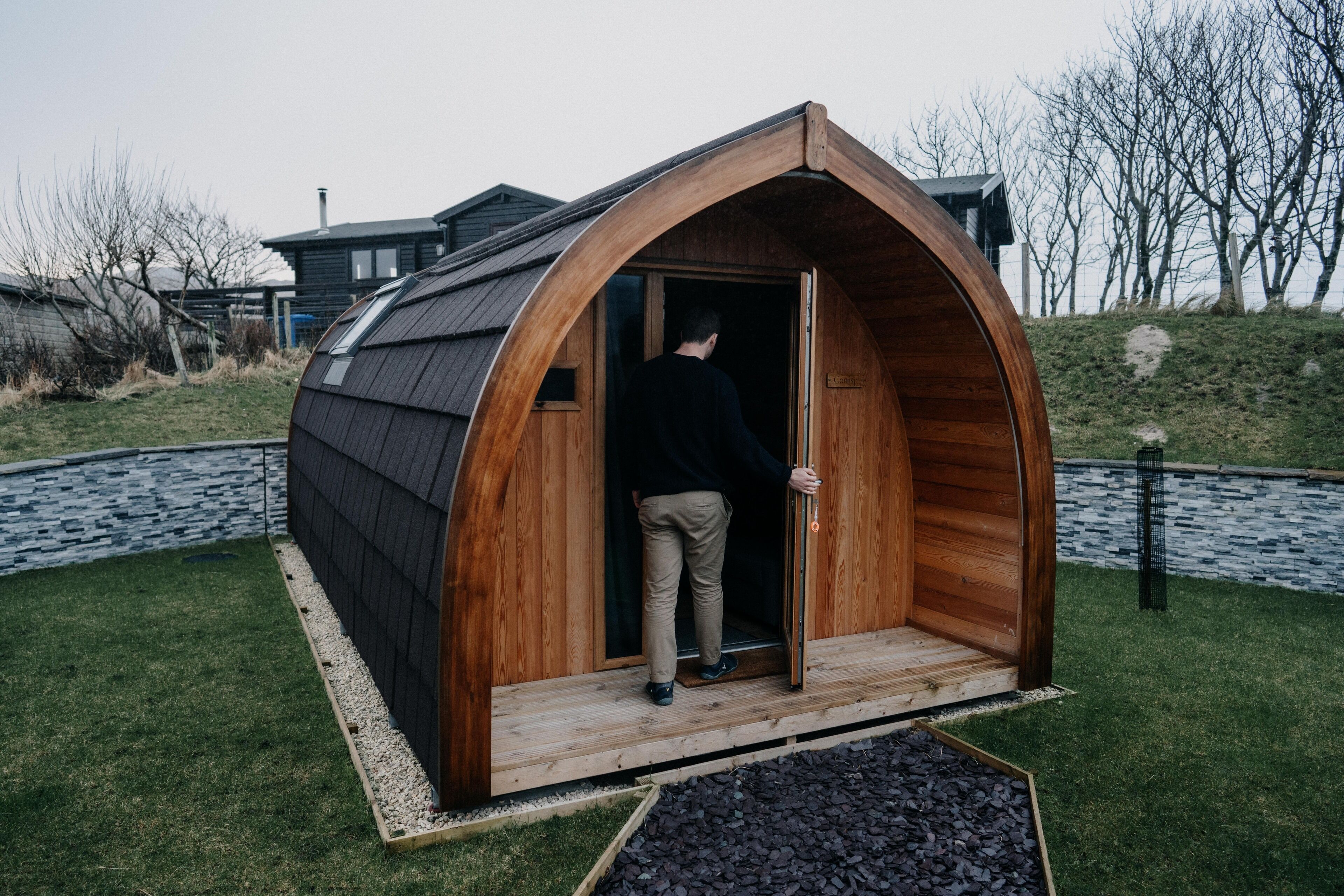 Een houten, halvemaanvormige constructie met een deur, een persoon die bij de ingang staat, en een grasveld met een pad van stenen en bomen op de achtergrond.