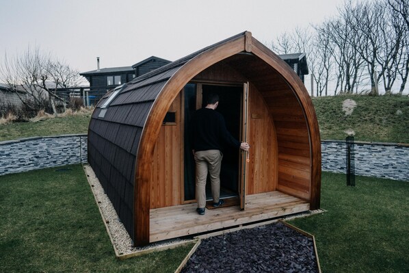 A wooden, semi-circular structure with a door, a person standing at the entrance, and a grassy area with a stone path and trees in the background. - North Coast 500 Pods (Achmelvich)
