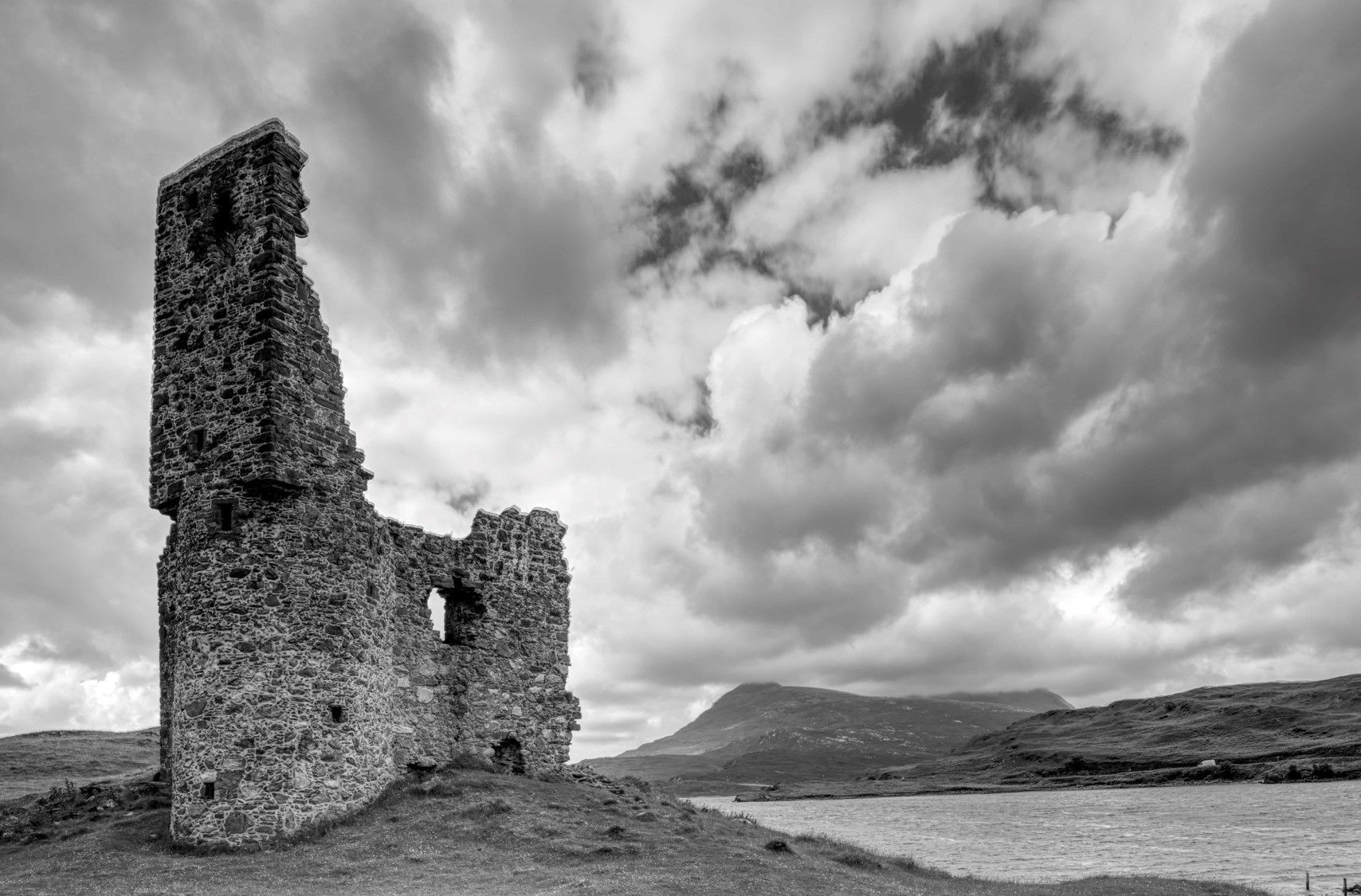 A historic stone tower with a large opening, surrounded by a hilly landscape and a body of water.