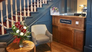 A hotel reception area with a wooden reception desk, a chair, a small table with a vase of flowers, and a staircase with white balusters. - Talardy, St Asaph by Marston’s Inns (Saint Asaph)