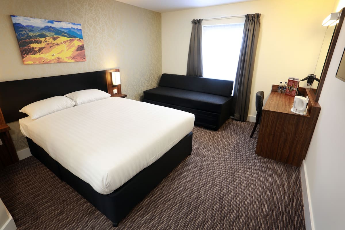 A hotel room with a large bed, a sofa, a desk with a mirror, and a window with curtains.