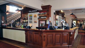 A bar area with a wooden counter, shelves of bottles, and a sign that reads "TALARDY". - Talardy, St Asaph by Marston’s Inns (Saint Asaph)