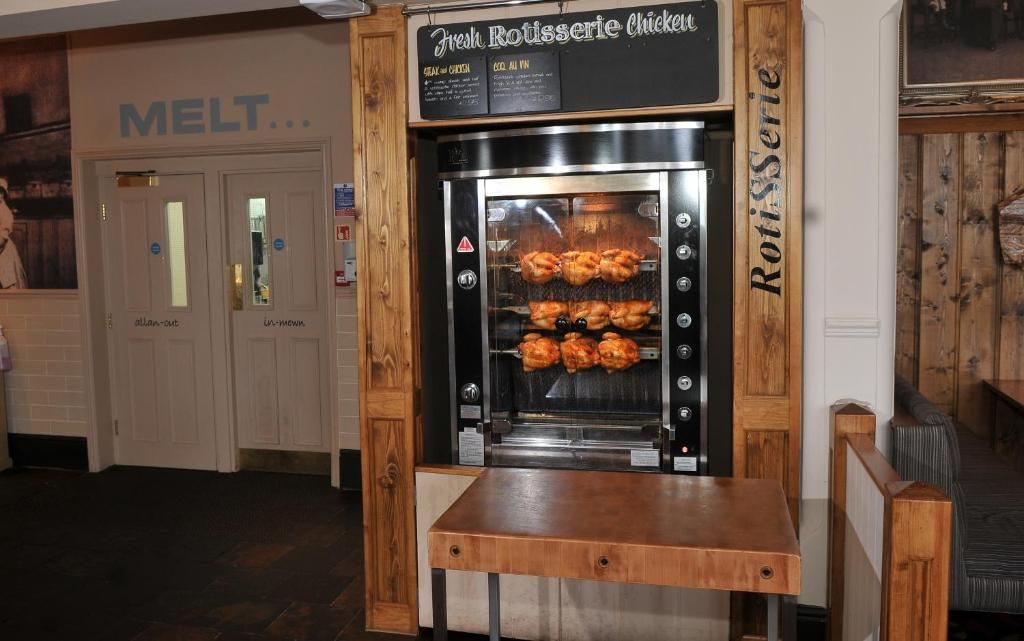 A commercial kitchen with a sign for 'Fresh Rotisserie Chicken' and a wooden table in front.