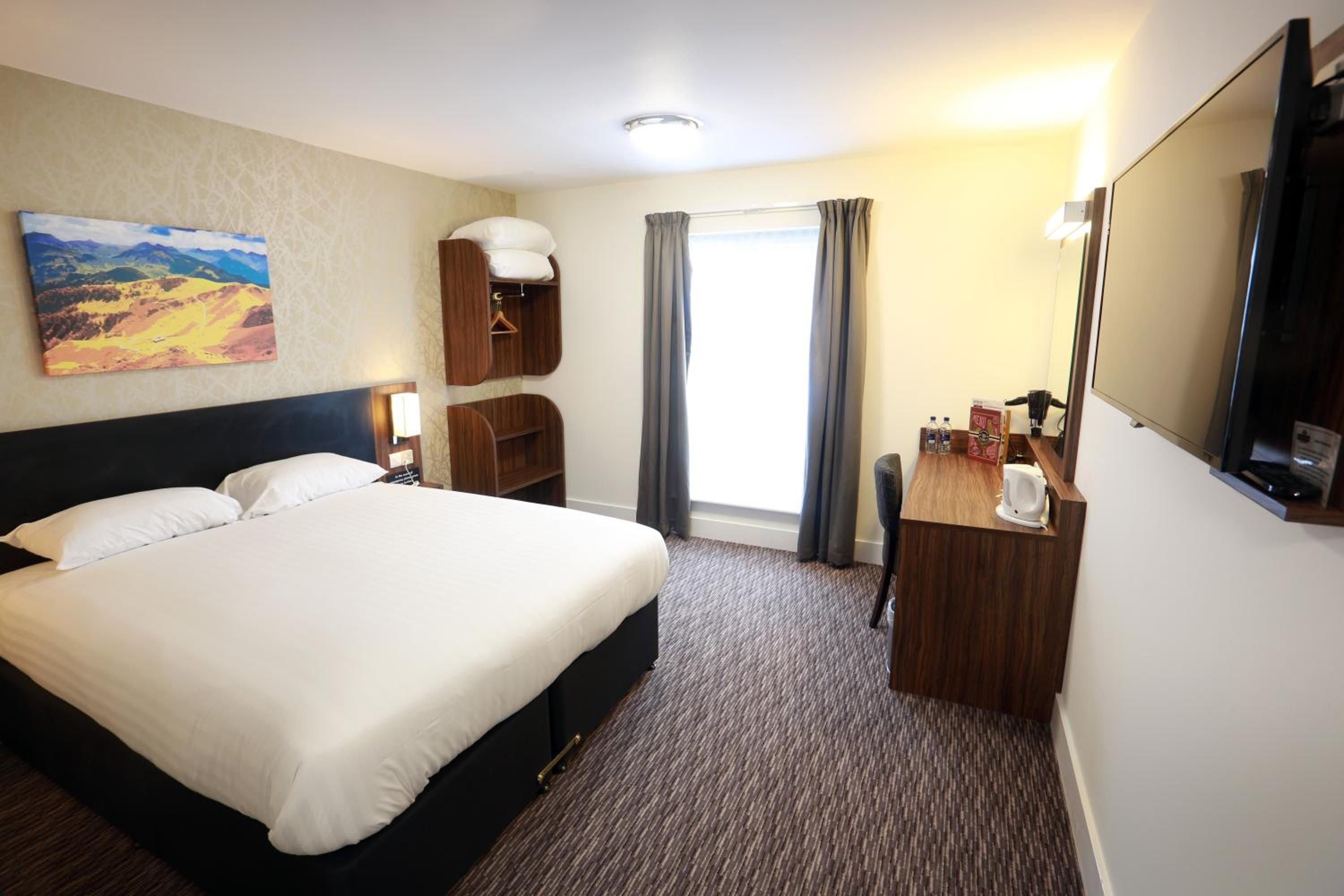Habitación de hotel con una cama grande, un escritorio con espejo, un pequeño espacio de descanso y un cuadro en la pared.