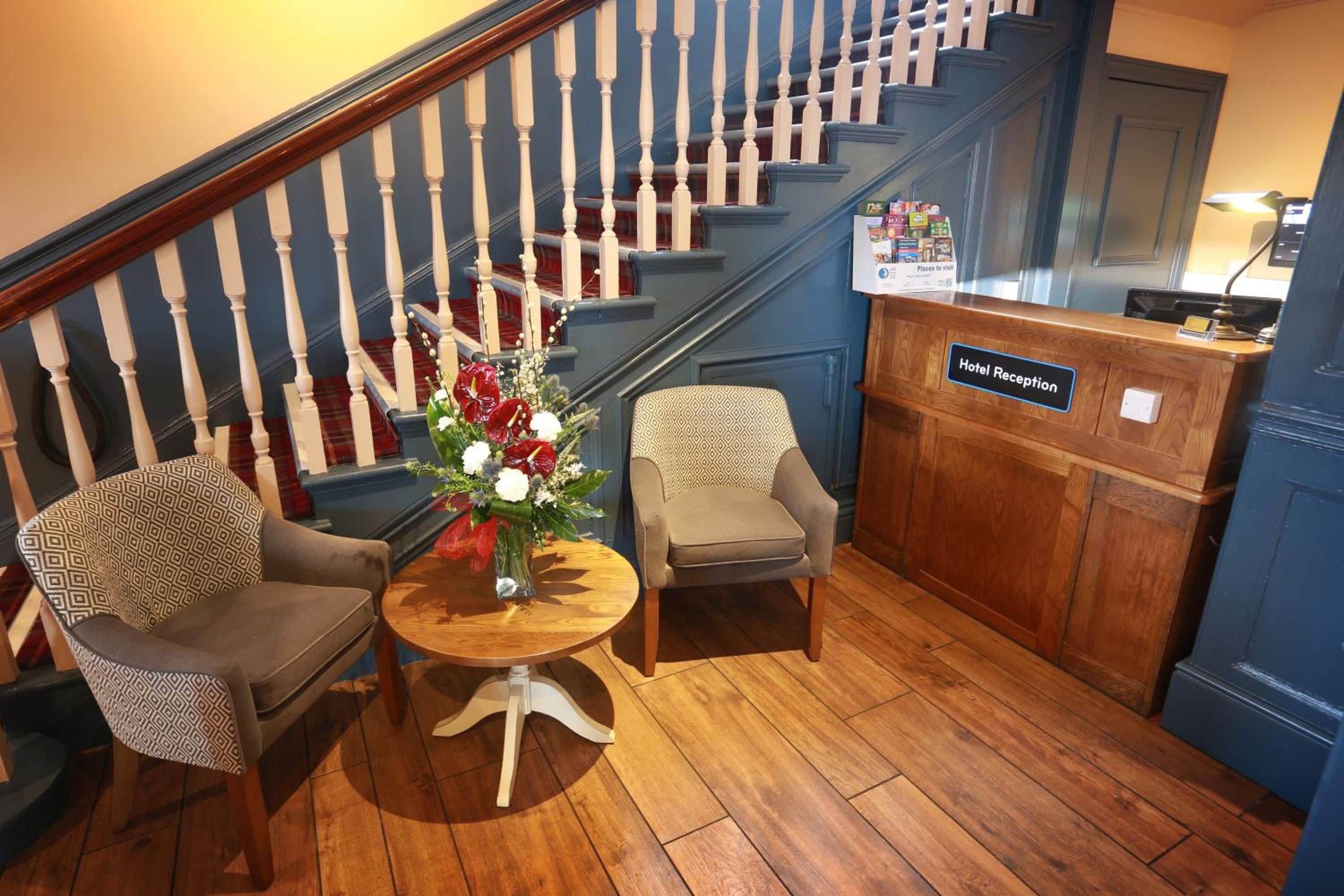A hotel reception area with a wooden floor, two patterned armchairs, a round table with a vase of flowers, and a staircase with white balusters.